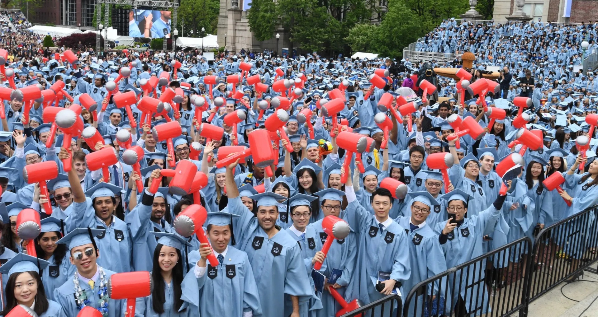 Hundreds of graduates in light blue robes and caps gathered in an outdoor university courtyard, enthusiastically holding red and silver foam hands in celebration, with historic academic buildings and green trees visible in the background.