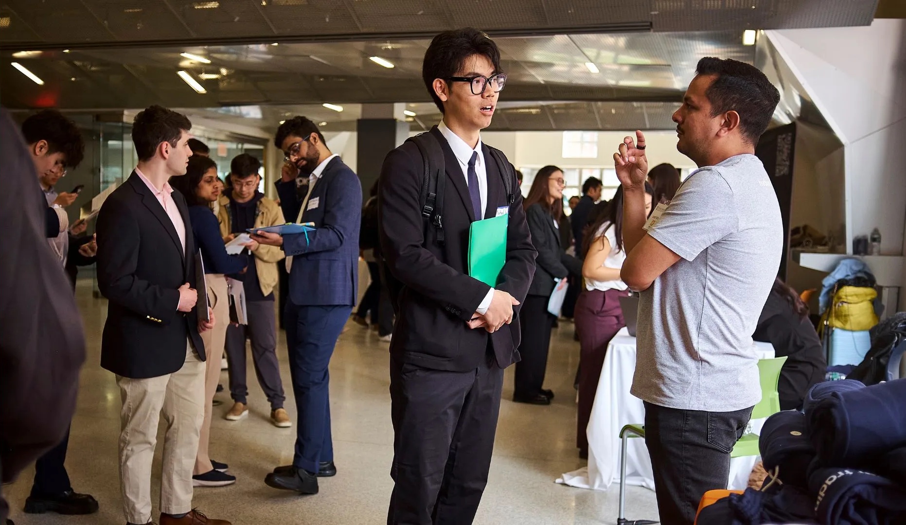 Large employer recruitment event in a modern glass-walled office corridor, with approximately 15 professionals in business attire engaged in networking conversations, one holding a green folder, gathered for what appears to be a career fair or recruitment event.