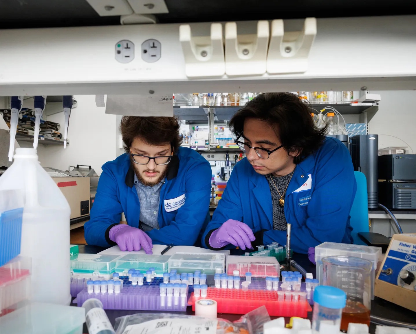Two students wearing blue lab coats and purple gloves examine sample containers with colored caps in a modern laboratory, surrounded by organized storage racks containing various lab materials and equipment on shelves in the background.