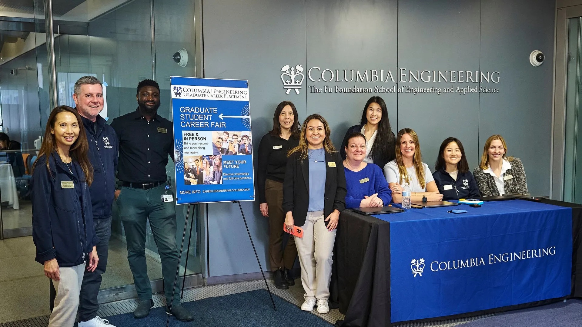 Ten Columbia Engineering staff and organizers posed together in front of a wall displaying the Columbia Engineering logo and 'The Fu Foundation School of Engineering and Applied Science,' with a blue 'Graduate Student Career Fair' sign visible, dressed in navy and professional attire.