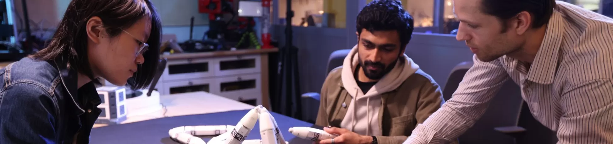 Three engineering students examine a white robotic hand prototype on a workbench in a lab.