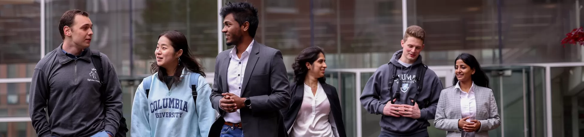 A group of graduate students walk and talk together on a campus walkway in front of a glass building, wearing a mix of casual Columbia sweatshirts and business attire.